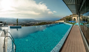 An infinity pool with clear blue water, a wooden deck, and a view of a distant city and hills.