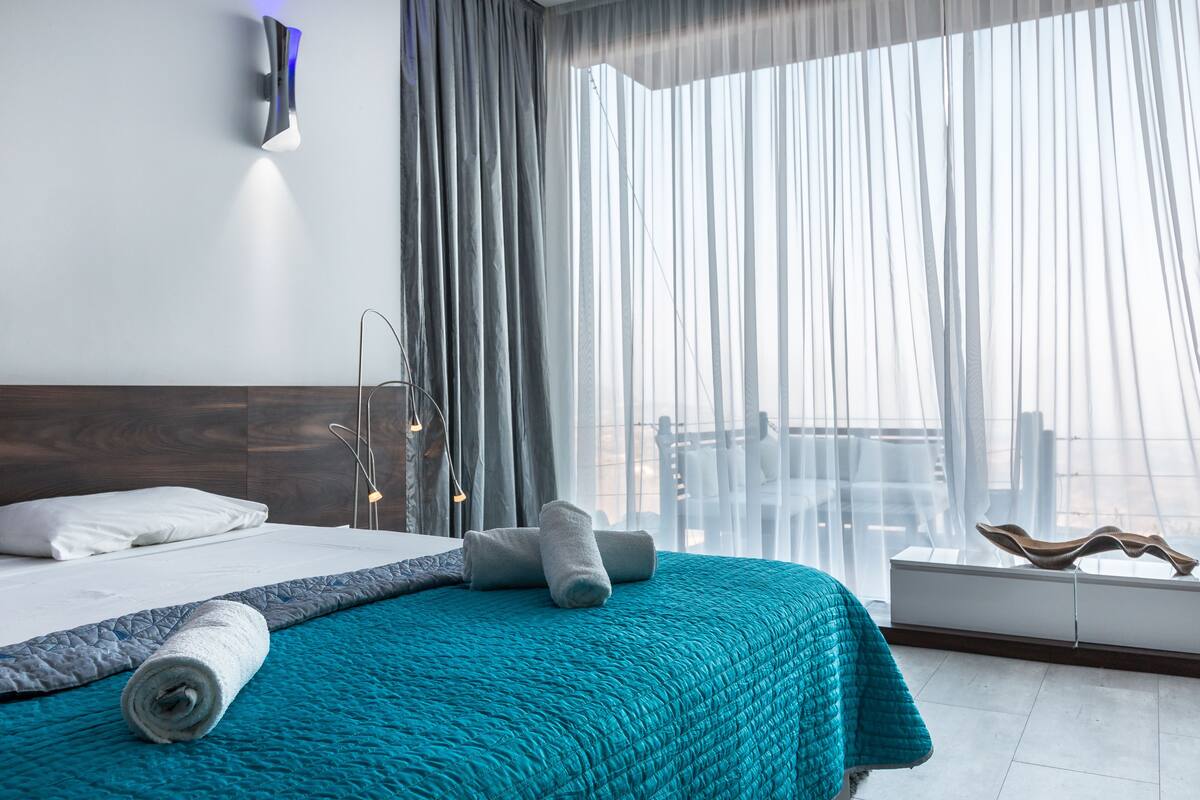 A modern hotel room with a large bed, a view of the city through sheer curtains, and a minimalist decor.