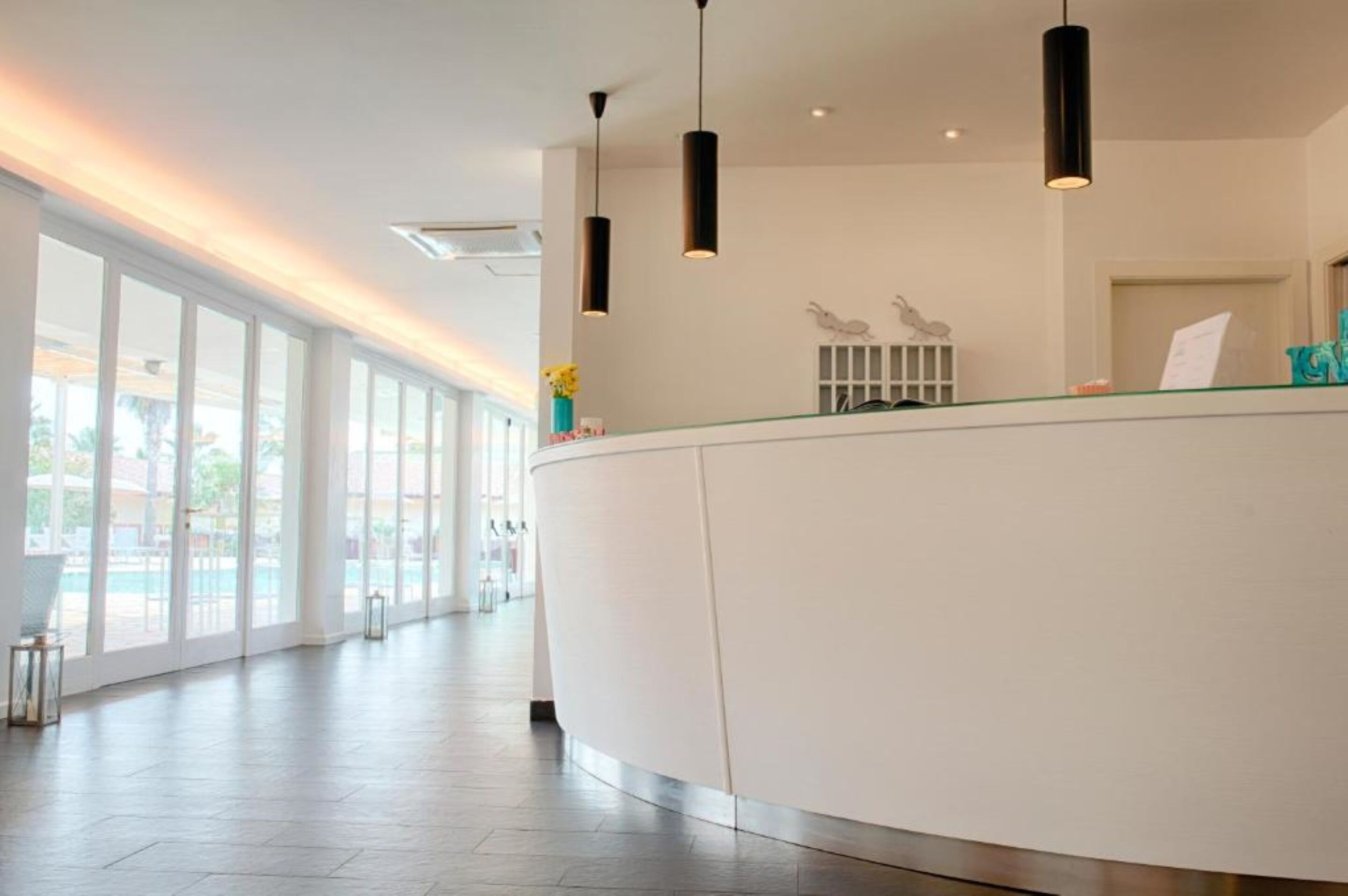 A modern hotel reception area with a curved reception desk, pendant lighting, and large windows offering views of the outdoor area.