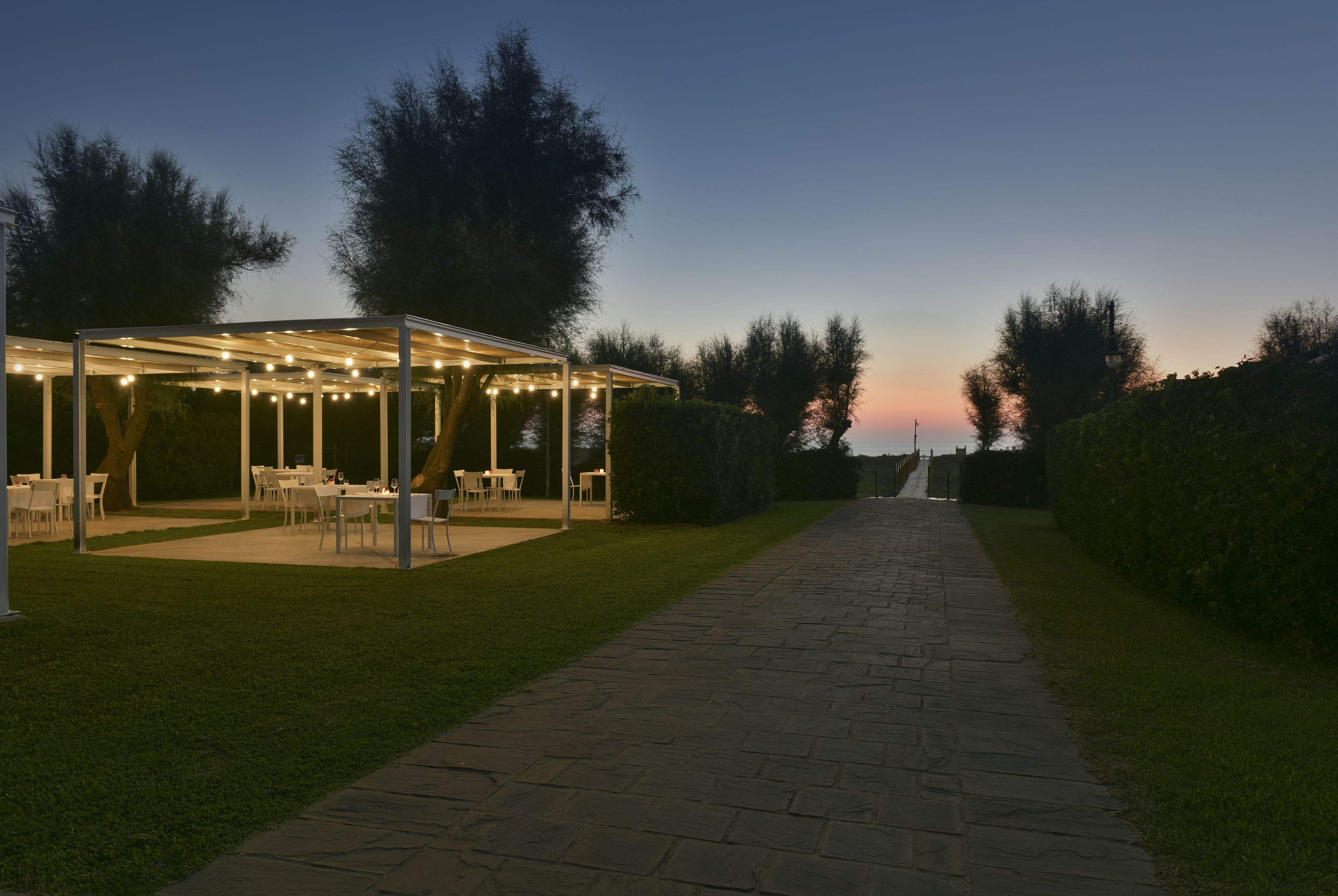 Un espace extérieur aménagé en terrasse avec des tables et des chaises, un coin salon couvert agrémenté de guirlandes lumineuses, et un sentier menant à un point de vue éloigné.
