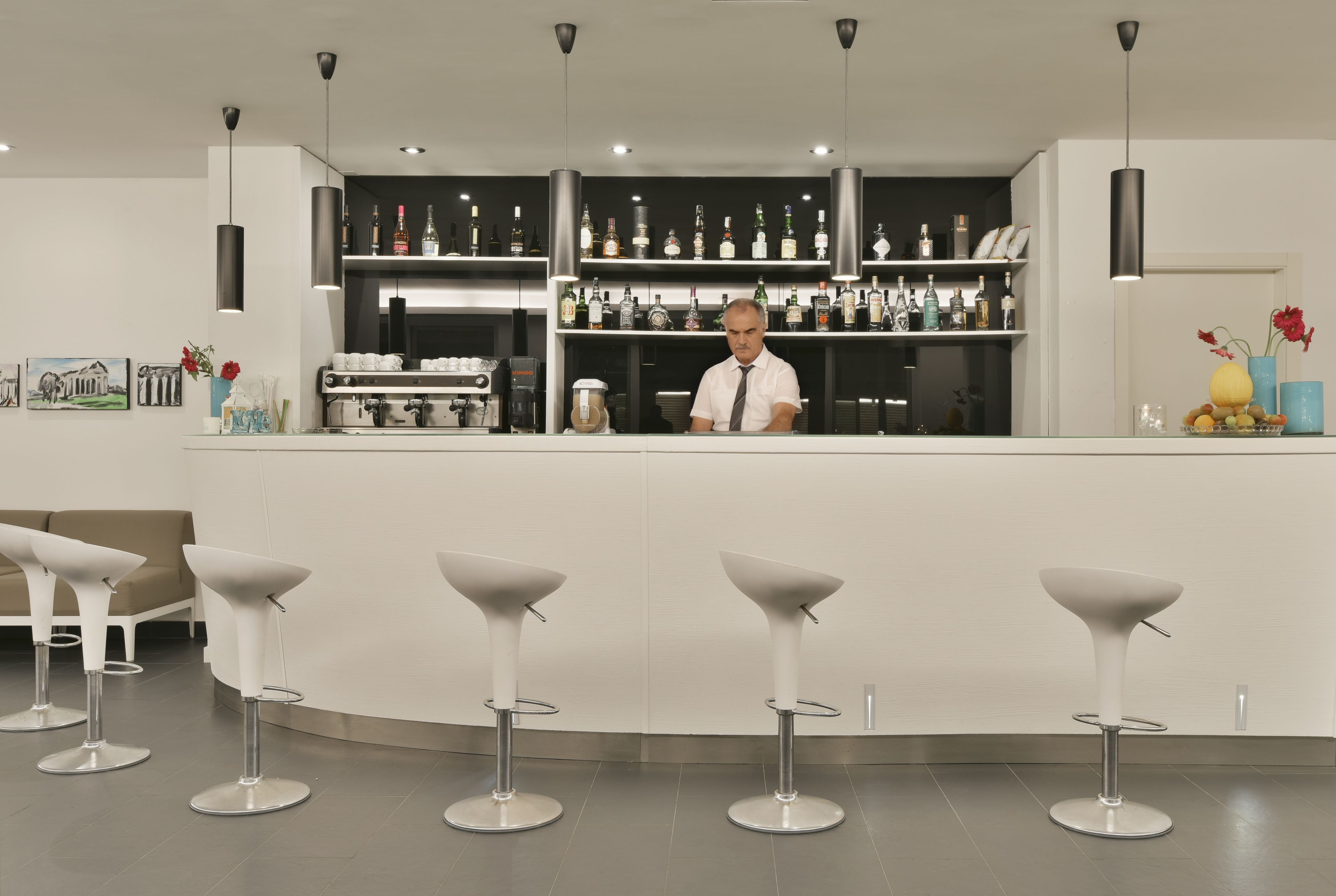 Eine Bar mit einem Barkeeper, weißen Barhocker und Regalen voller Flaschen und Gläsern.