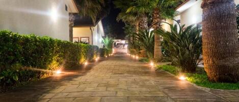 A nighttime pathway flanked by palm trees and lit by small lights, leading to a building with a stone facade.