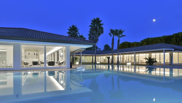 A modern pool area with clear glass walls, a well-lit interior, and palm trees in the background. - Paestum Inn Beach Resort (Capaccio-Paestum)