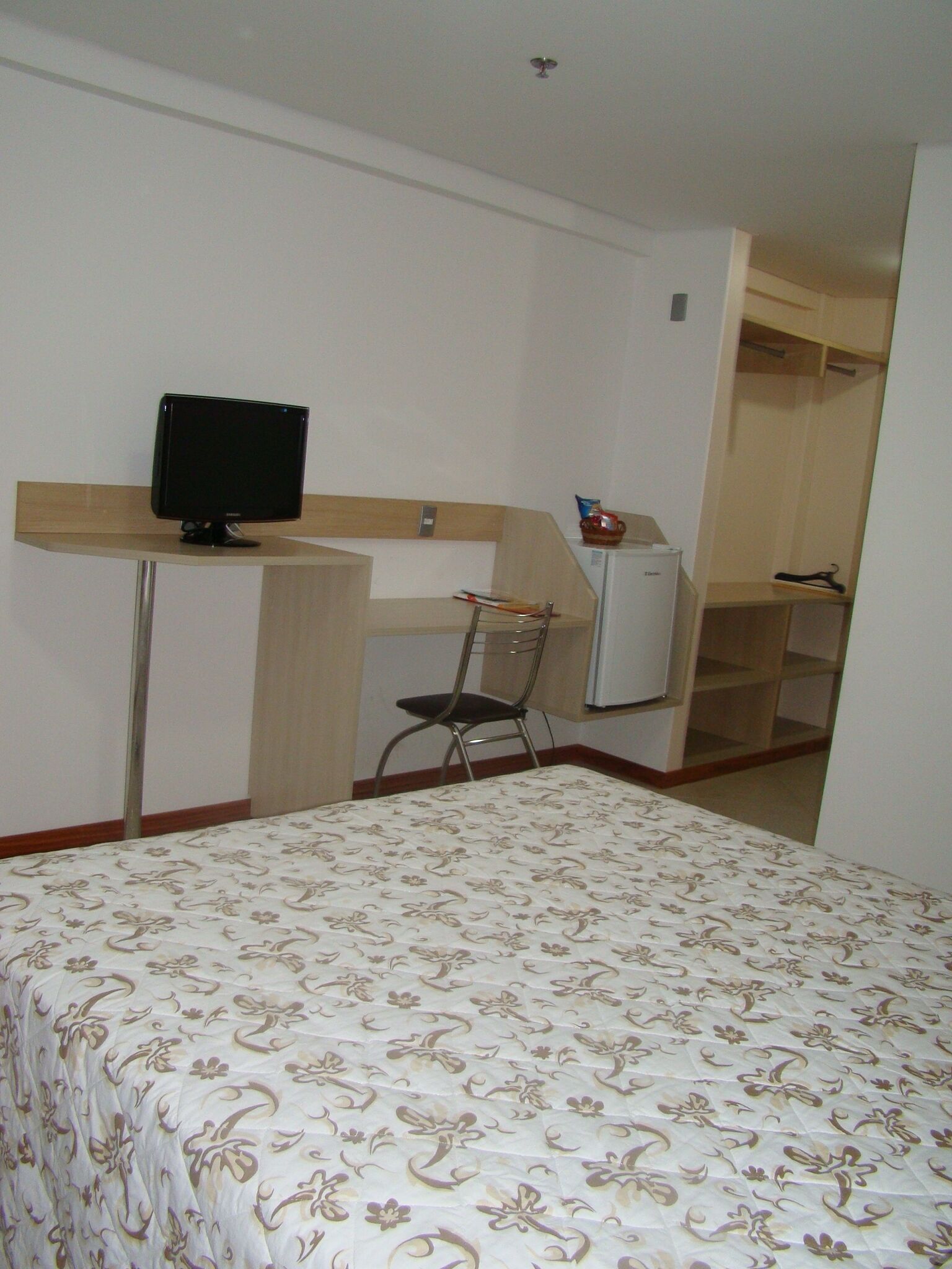 Quarto de hotel com uma cama, escrivaninha com computador, cadeira e guarda-roupa.