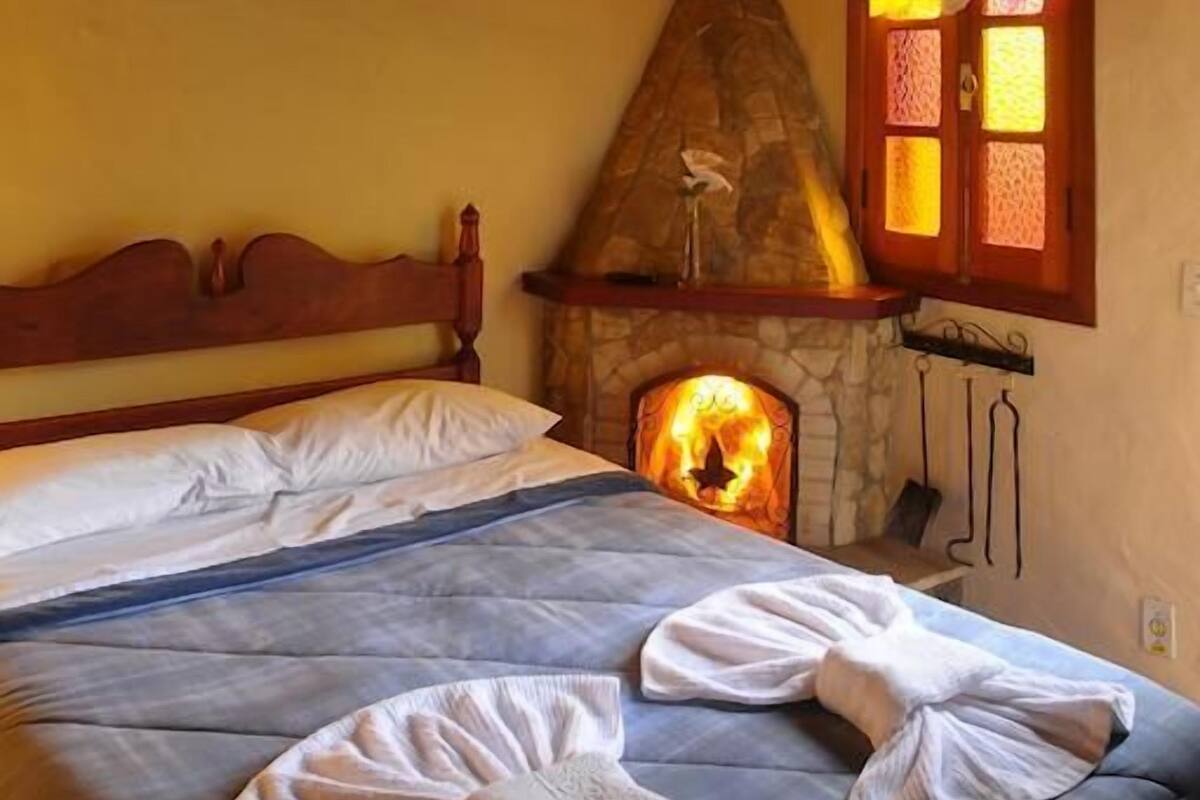 Un dormitorio con chimenea de piedra, vigas de madera, una cama con ropa blanca y una ventana con cortinas estampadas.