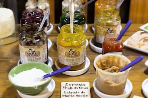Uma mesa com potes de doce 'Chale da Tânia', uma tigela com um produto branco e um pote de manteiga de amendoim.