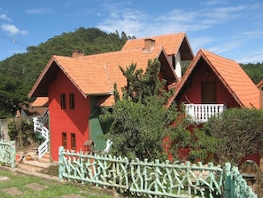A traditional house with a red facade and a tiled roof, surrounded by greenery and a white fence.