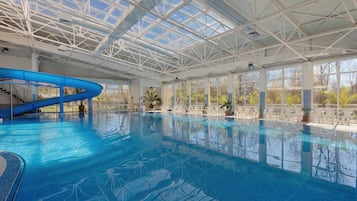 An indoor swimming pool with a glass ceiling, a blue slide, and potted plants around the pool area.