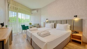 A hotel room with two beds, a desk, a chair, a lamp, and a view of greenery outside. - Augusta SPA hotel (Hisarja)