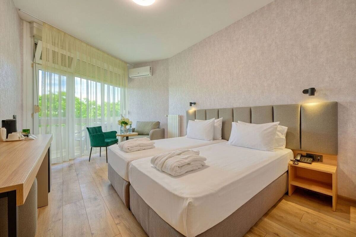 A hotel room with two beds, a desk, a chair, a lamp, and a view of greenery outside.
