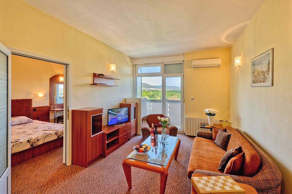 A hotel room with a bed, a TV, a dining table with chairs, a sofa, and a view of the outdoors.