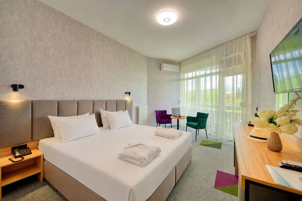 A hotel room with a large bed, bedside tables, a desk with a chair, a TV, and a window with curtains.