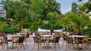 An outdoor patio area with round tables and wicker chairs, surrounded by lush greenery and a boat in the background. - Augusta SPA hotel (Hisarja)