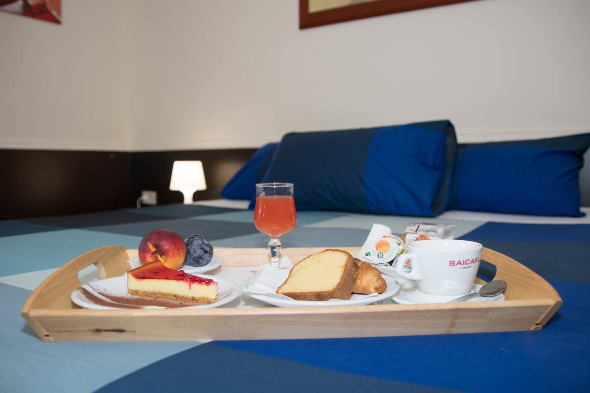 A breakfast tray with a slice of cheesecake, a peach, a glass of juice, a slice of bread, a muffin, and a cup labeled "SAICAR" on a bed with blue bedding and pillows.