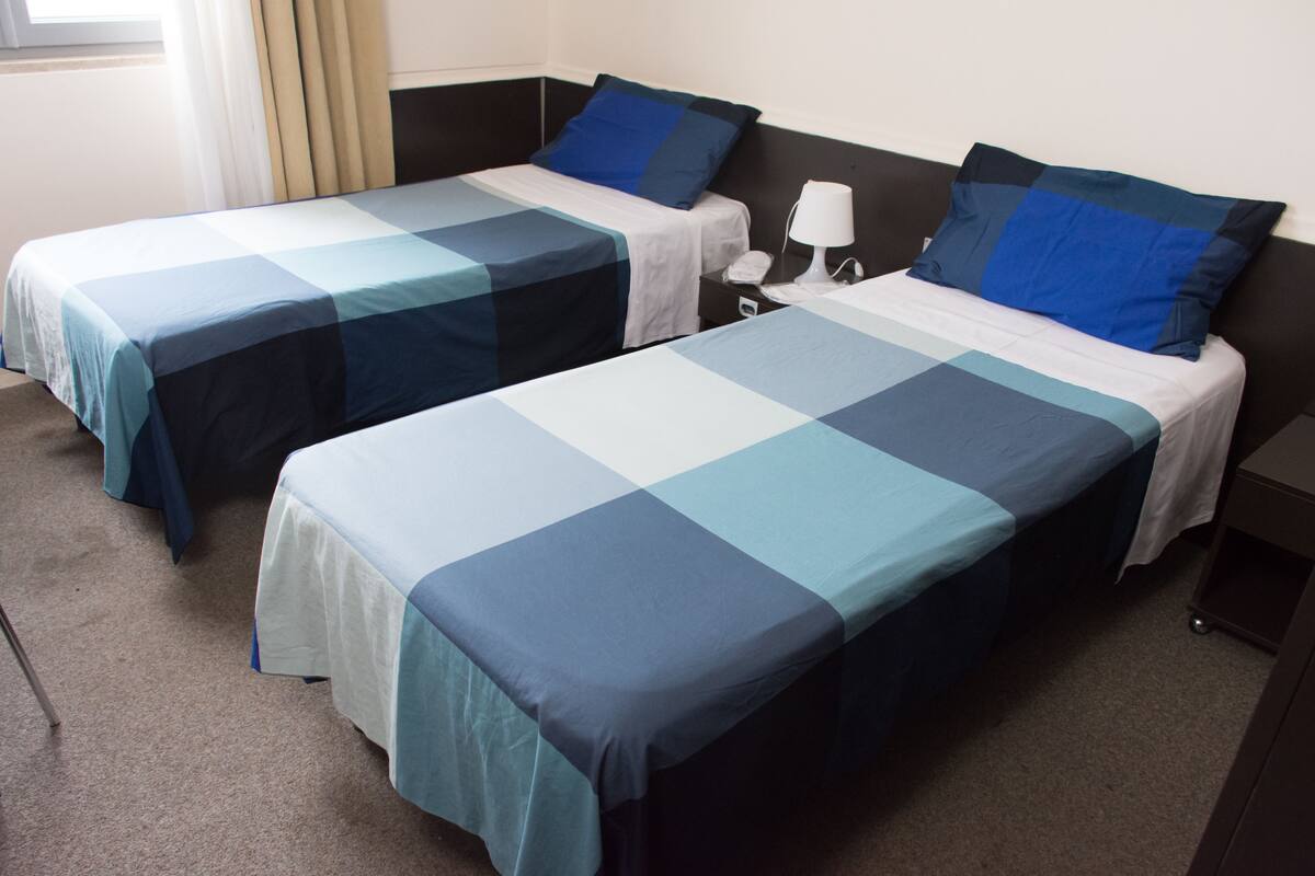 Two single beds with blue and white bedding, a bedside table with a lamp, and a window with curtains.