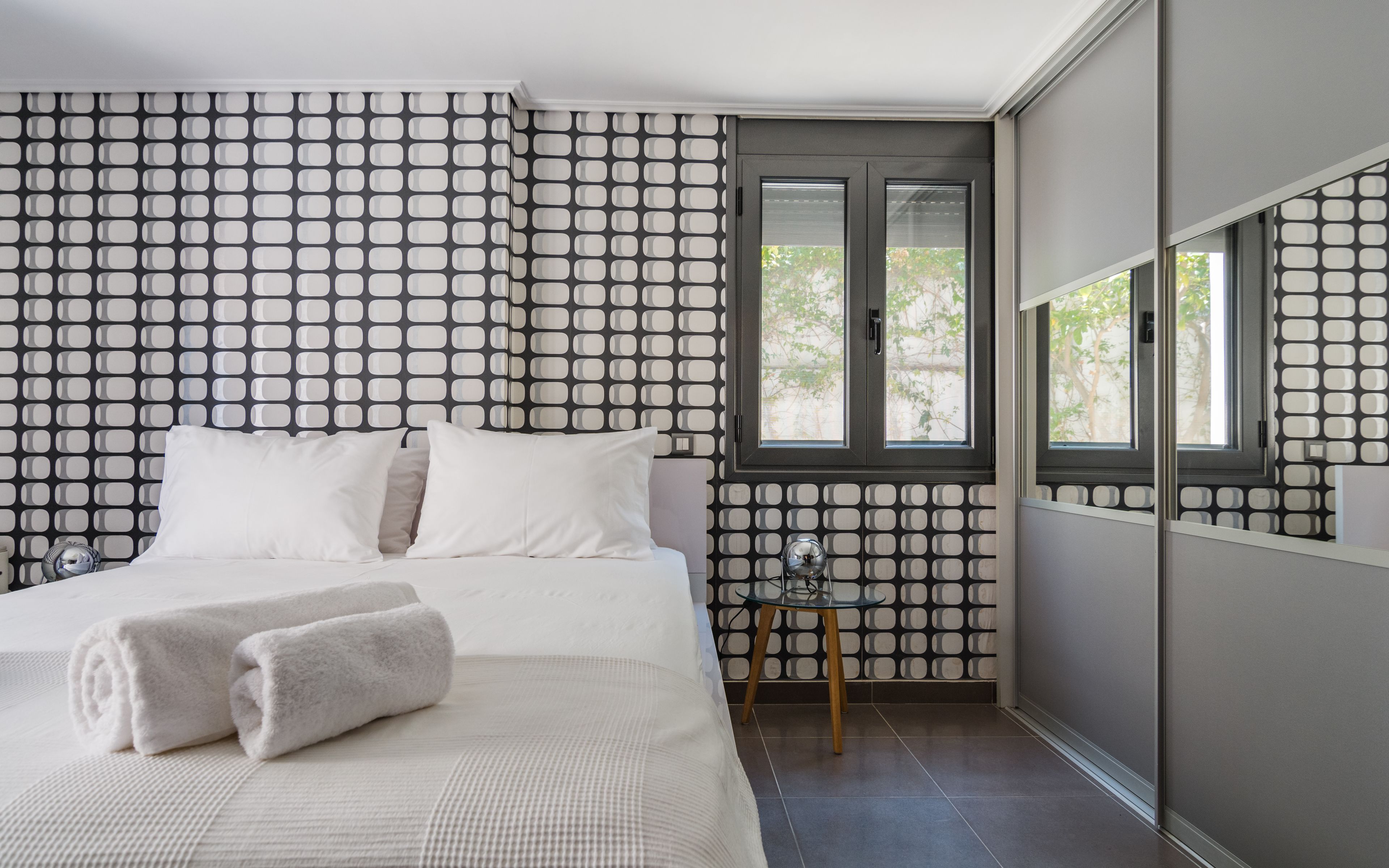 A bedroom with a bed, two pillows, a bedside table with a lamp, and a window with a view of trees.