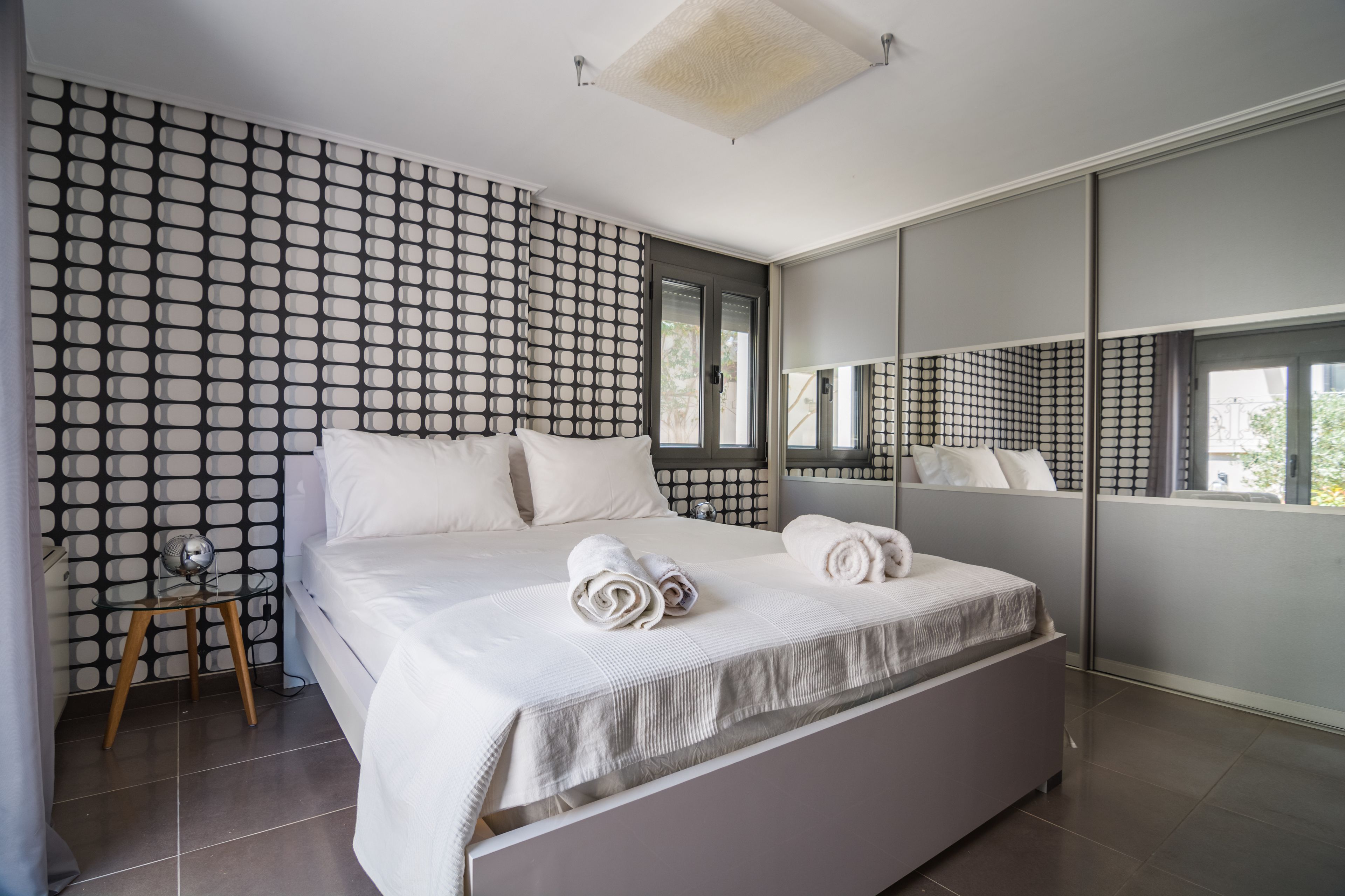 Une chambre moderne avec un grand lit, une petite table de chevet, un miroir et un mur décoré d’un motif.