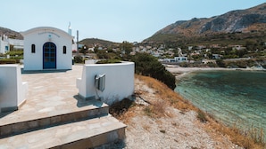Un edificio blanco con una puerta azul, una escalera de piedra que lo conduce, y una vista despejada de una zona costera con casas y montañas al fondo.