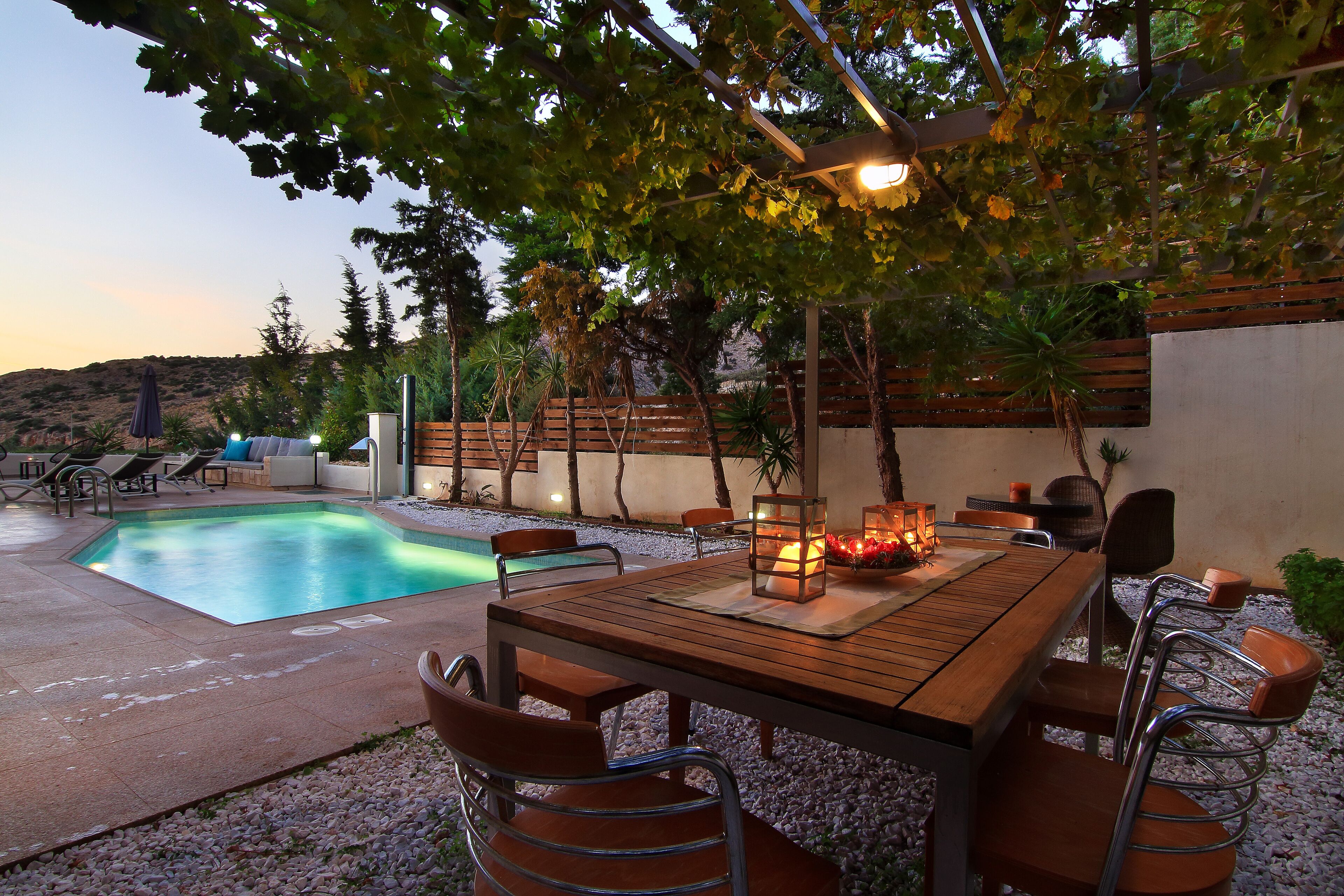 Zona piscina con tavolo e sedie in legno, circondata da un giardino alberato e vista su un paesaggio collinare.