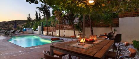 Zona piscina con tavolo e sedie in legno, circondata da un giardino alberato e vista su un paesaggio collinare.