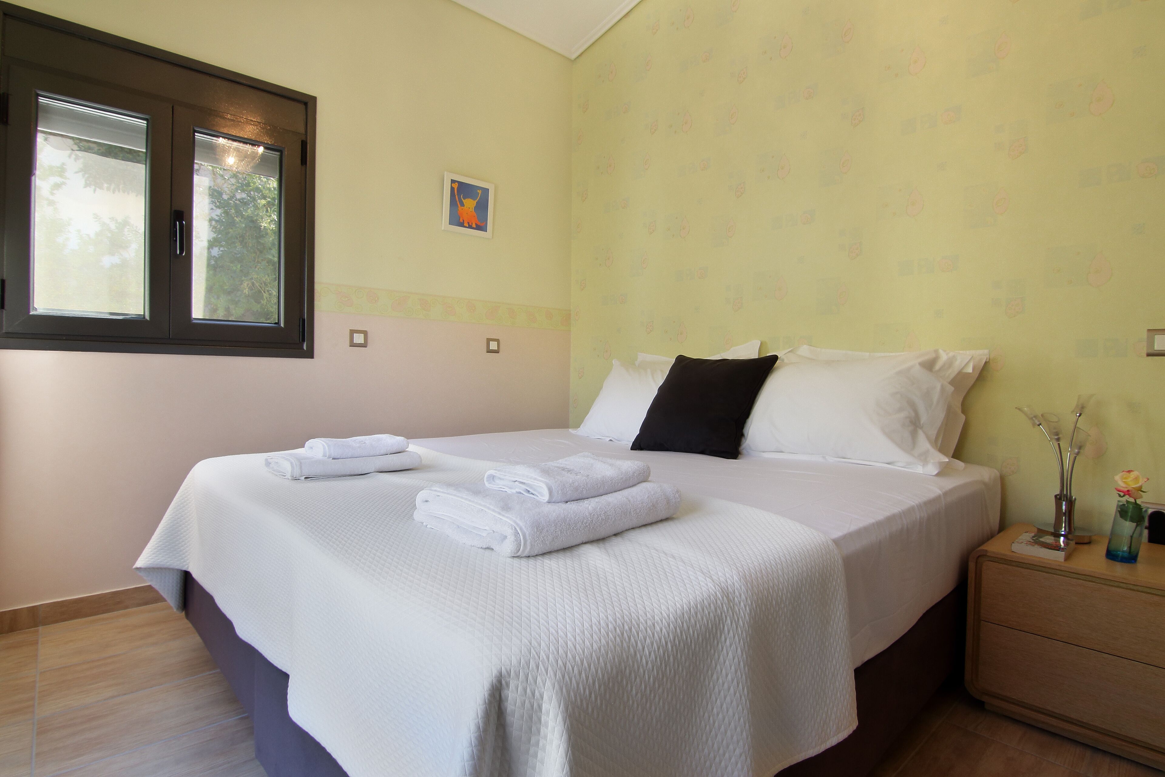 A neatly made bed with white linens and towels, a bedside table with a vase and a flower, and a window with blinds.