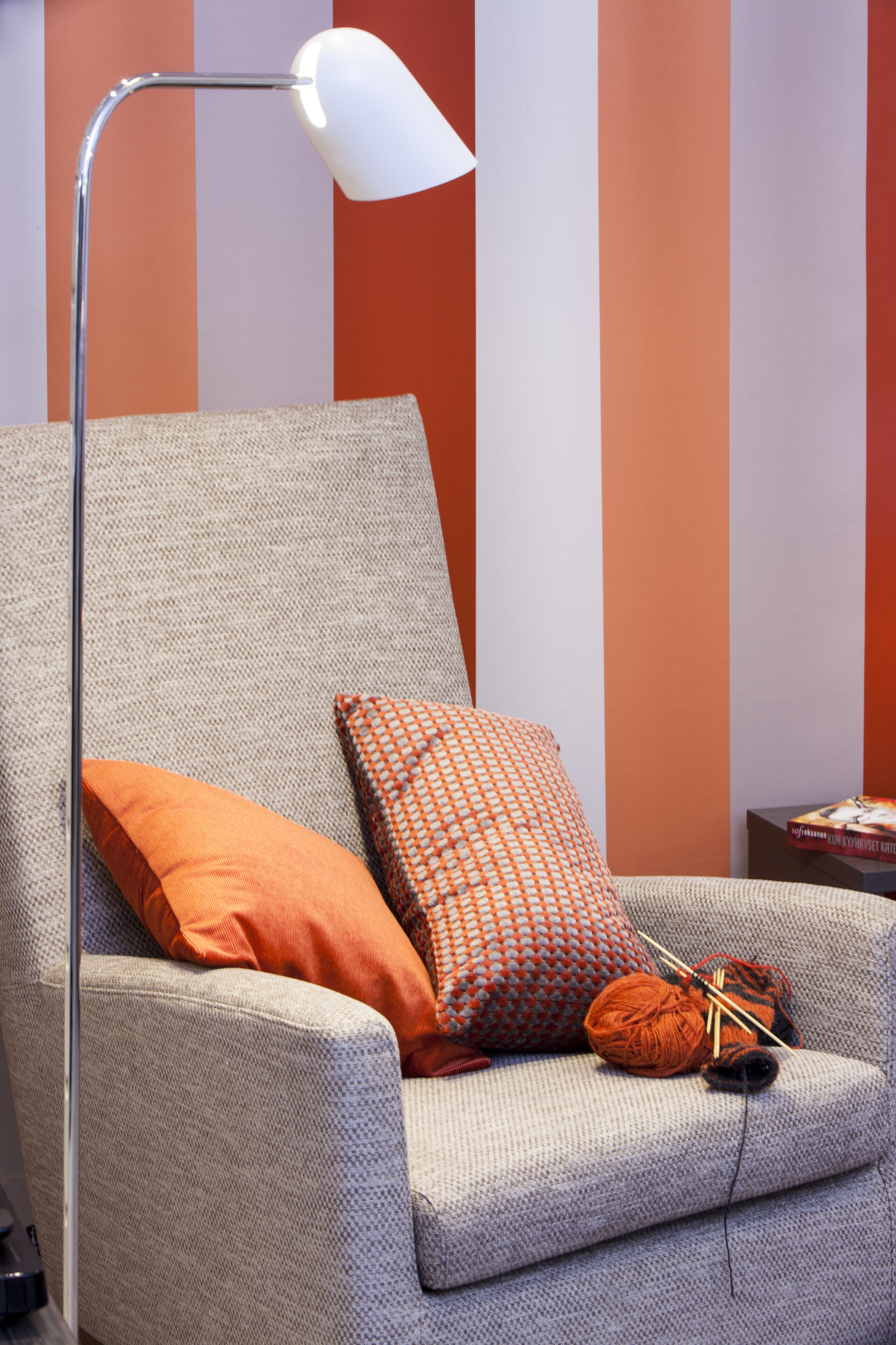 A grey armchair with orange and patterned cushions, a floor lamp, and a small table with a book.