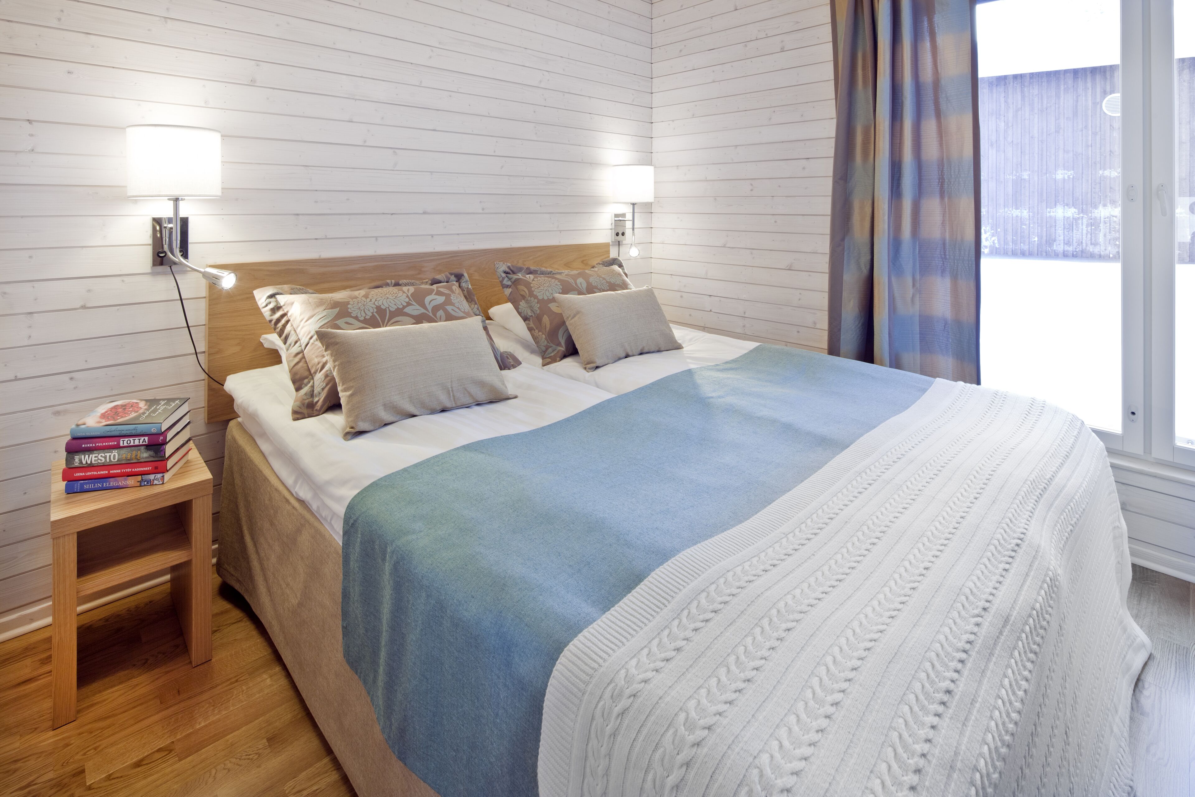 Een net opgemaakt bed met een blauw-witte deken, twee wandlampen om te lezen, een houten nachtkastje met boeken en een raam met gordijnen.