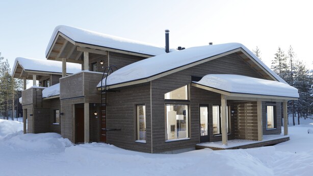 A modern two-story house with a sloped roof covered in snow, large windows, and a wooden exterior.