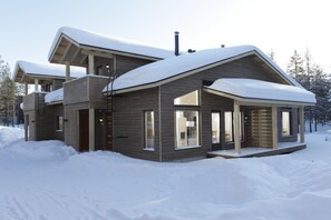 A modern two-story house with a sloped roof covered in snow, large windows, and a wooden exterior.