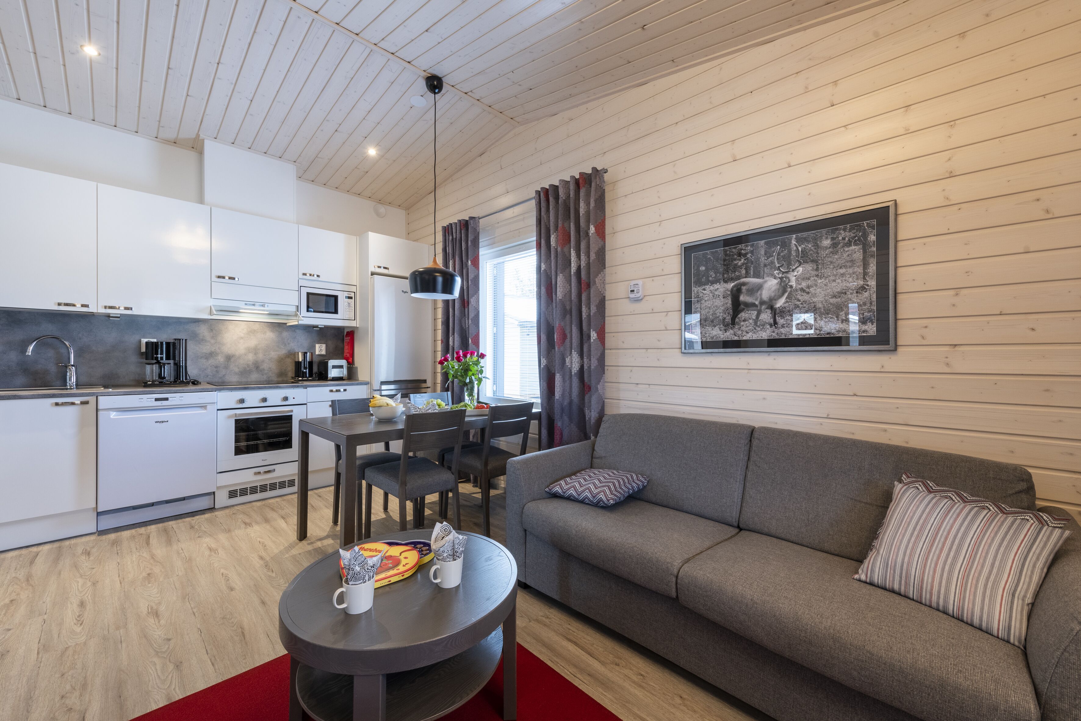 A modern kitchen with white cabinets and stainless steel appliances, a dining area with a table and chairs, and a gray sofa with patterned pillows.
