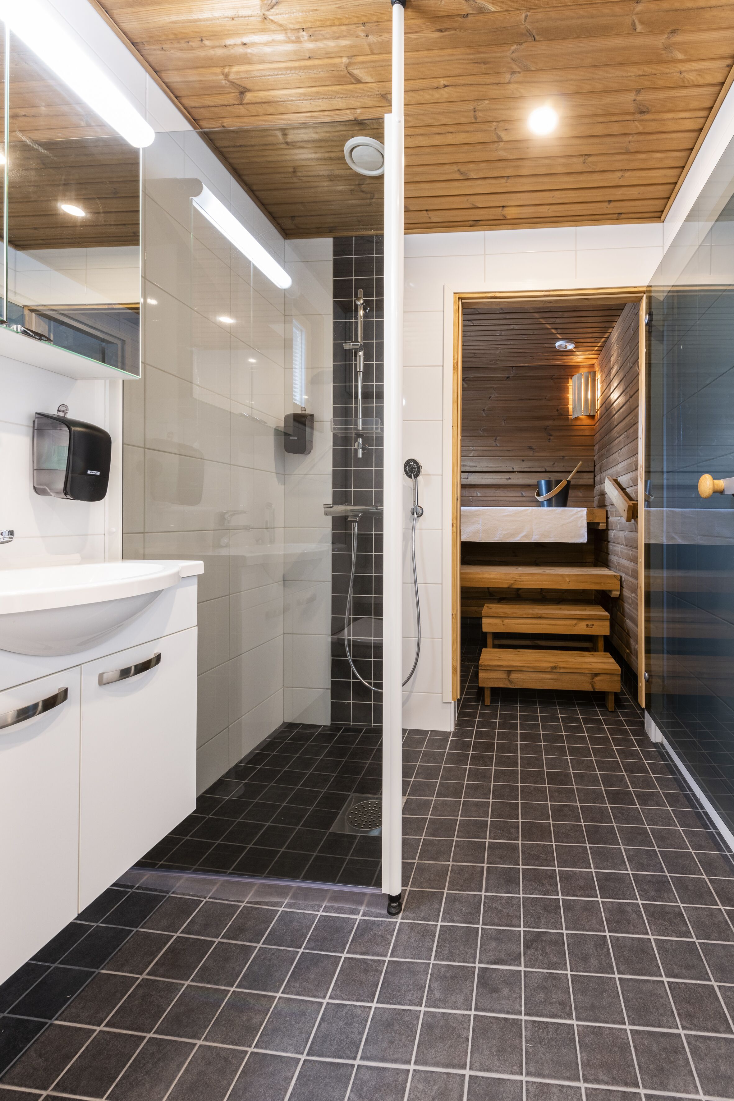 A modern bathroom with a wooden ceiling, a glass shower enclosure, a white sink, and a small wooden bench.