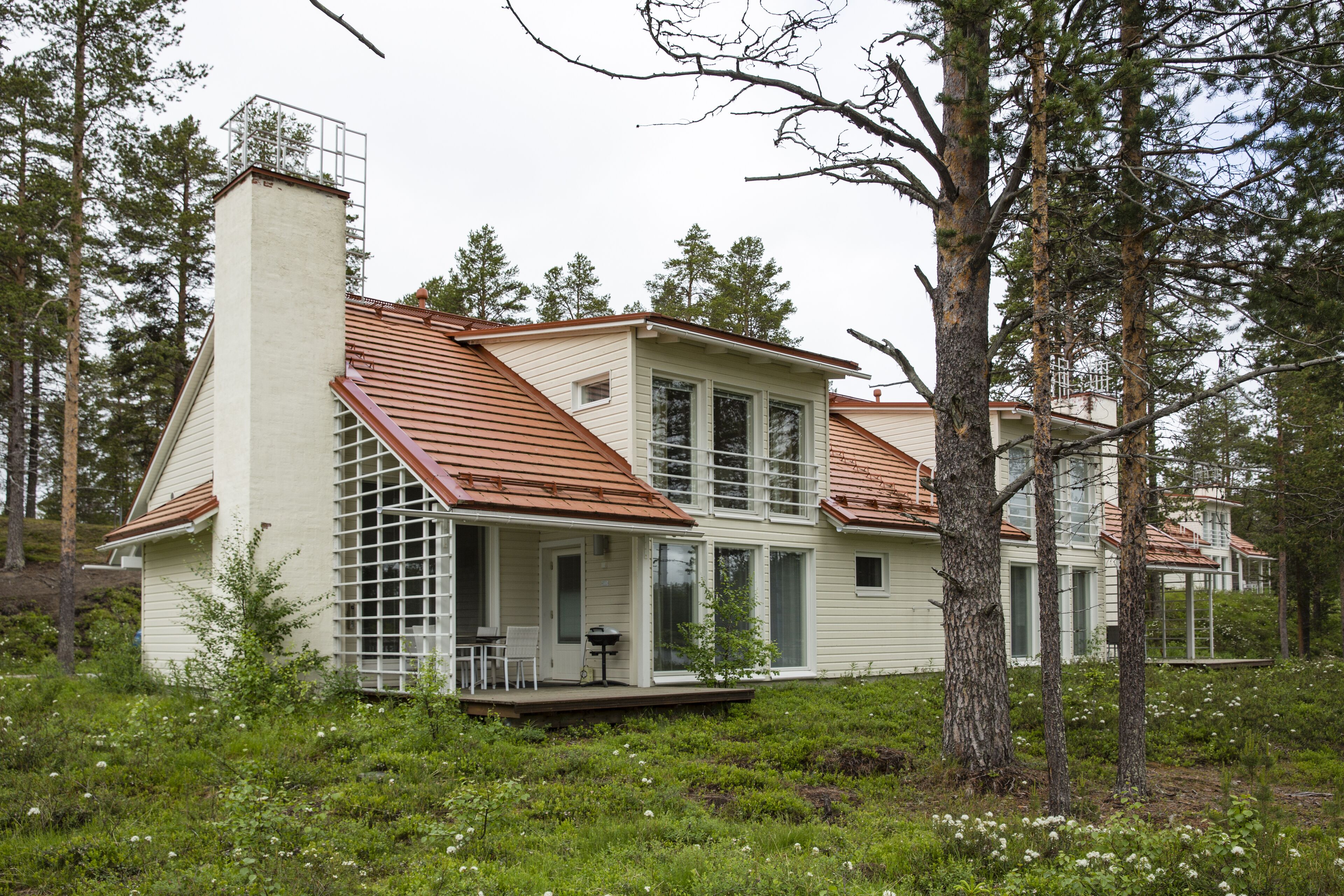 Una casa de dos pisos con techo inclinado, chimenea y porche con mobiliario exterior.