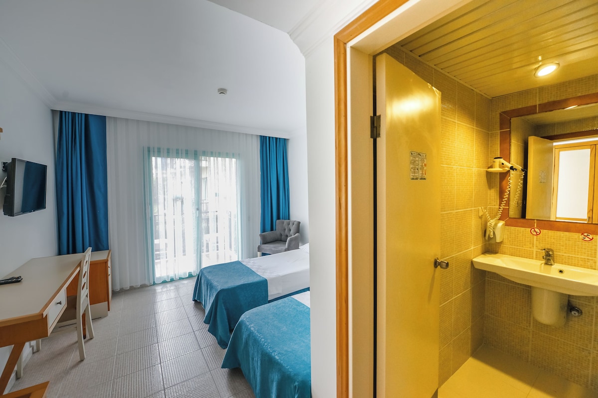 A hotel room with a desk, two beds, a chair, and a bathroom with a sink and mirror.