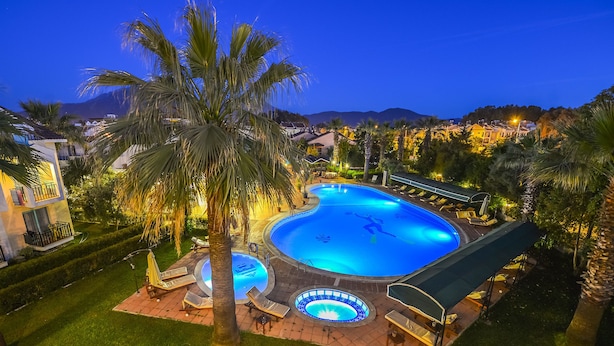 A swimming pool with a slide, surrounded by palm trees and lounge chairs, set against a mountainous backdrop. - Rebin Beach Hotel (Fethiye)