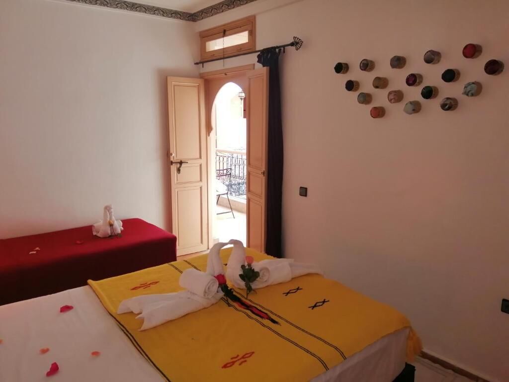 A hotel room with two beds, one with a yellow bedspread and the other with a red bedspread. There is a small table with a red cloth and a white decorative item. A wall-mounted clock and a series of small decorative items are visible.