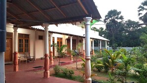 A covered outdoor area with columns and a tiled floor, surrounded by greenery and a white fence. - Hotel Alas Garden (Trincomalee)