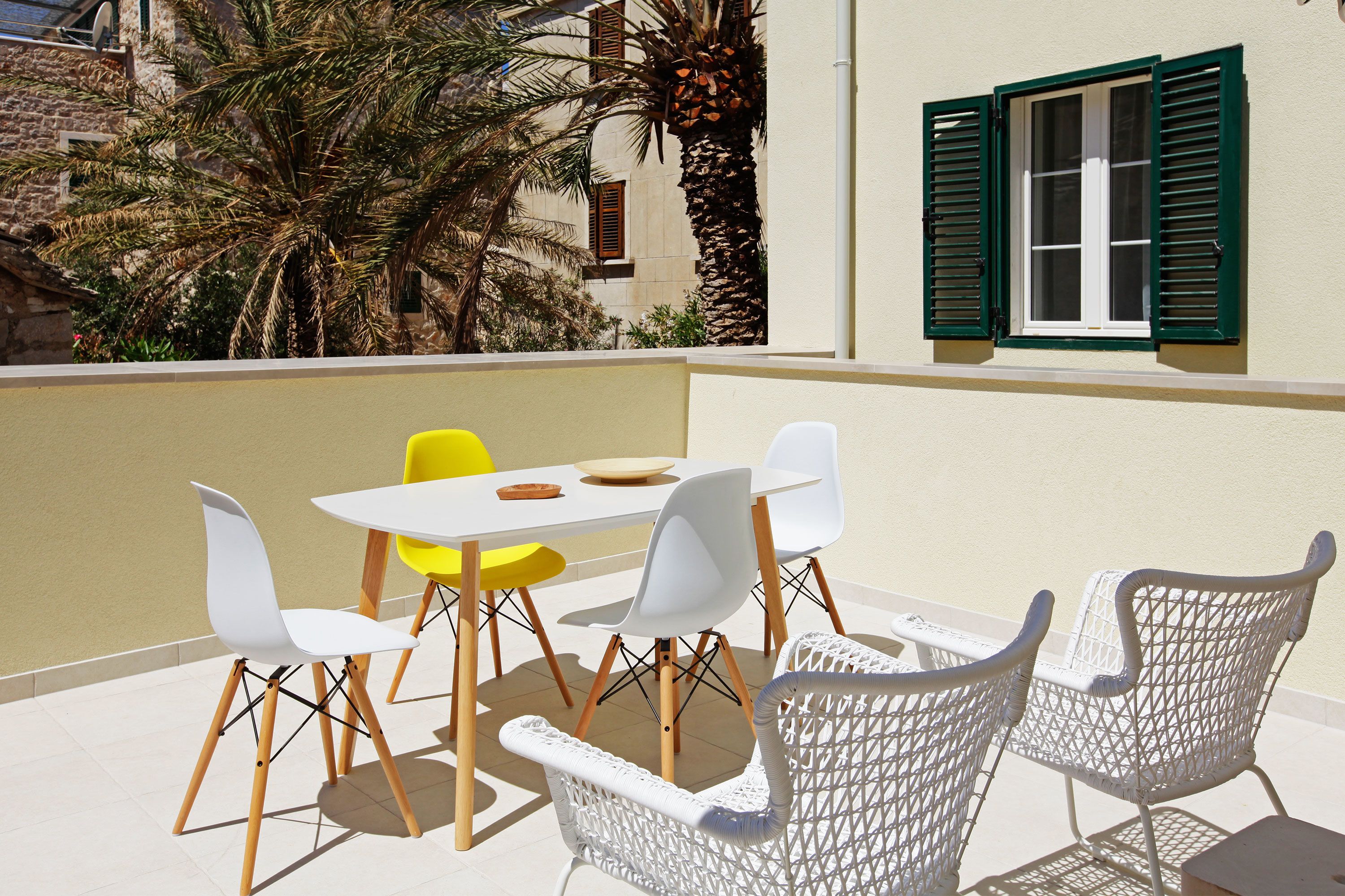 A patio with a table and chairs, a window with green shutters, and a palm tree.