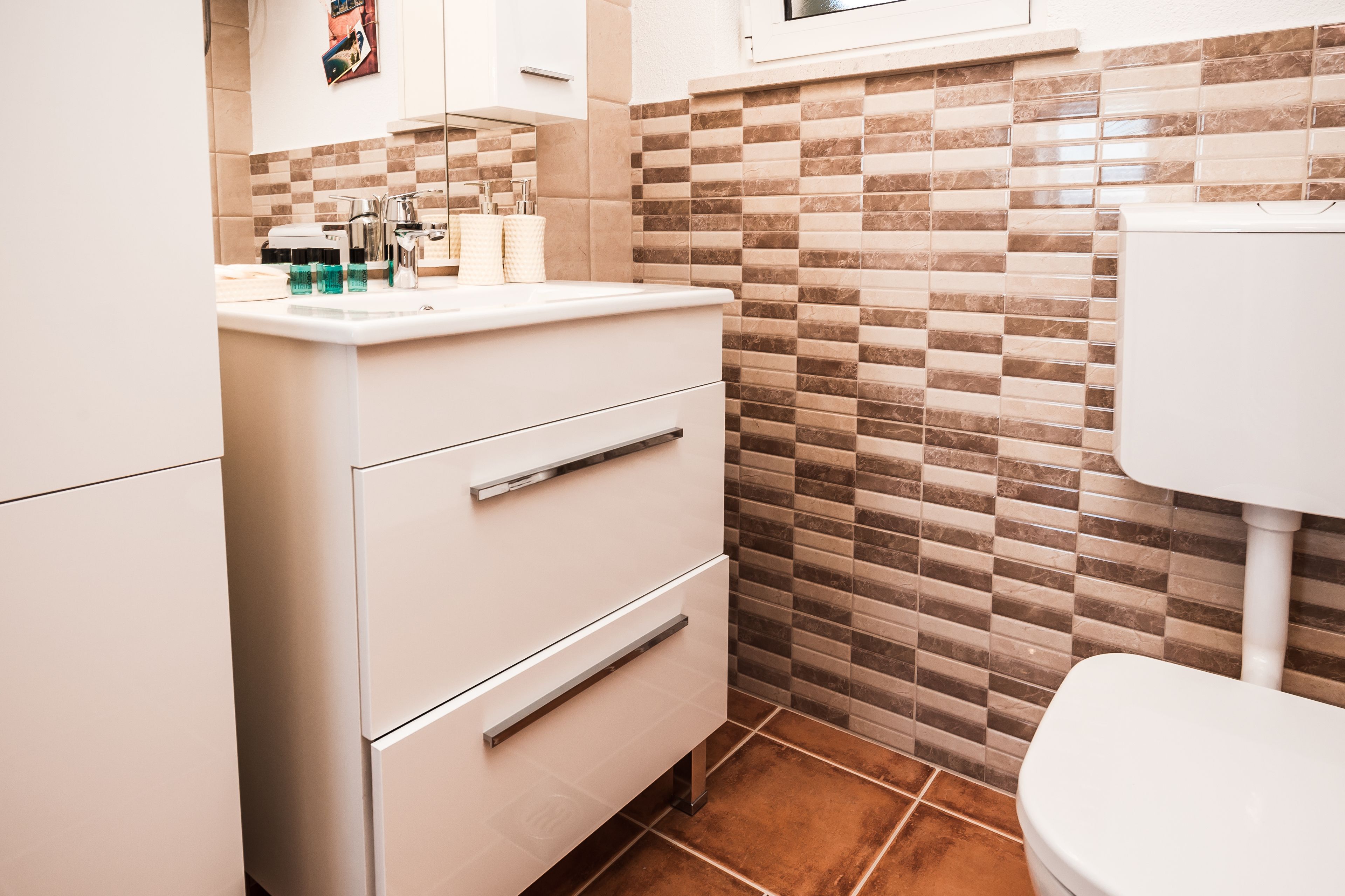 A modern bathroom with a white sink, two drawers, a mirror, and a toilet. The walls have a checkered tile pattern.