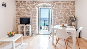 A room with a stone wall, a balcony with a view of the sea, a TV on a stand, a dining table with chairs, and a small white shelf with a fruit bowl. - Luxury Seaside Apartments (Bol)