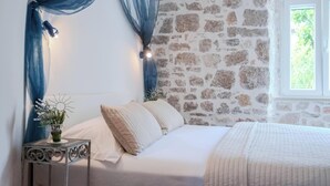 A bedroom with a stone wall, a bed with white bedding, a bedside table with a vase of flowers, and blue drapes. - Luxury Seaside Apartments (Bol)