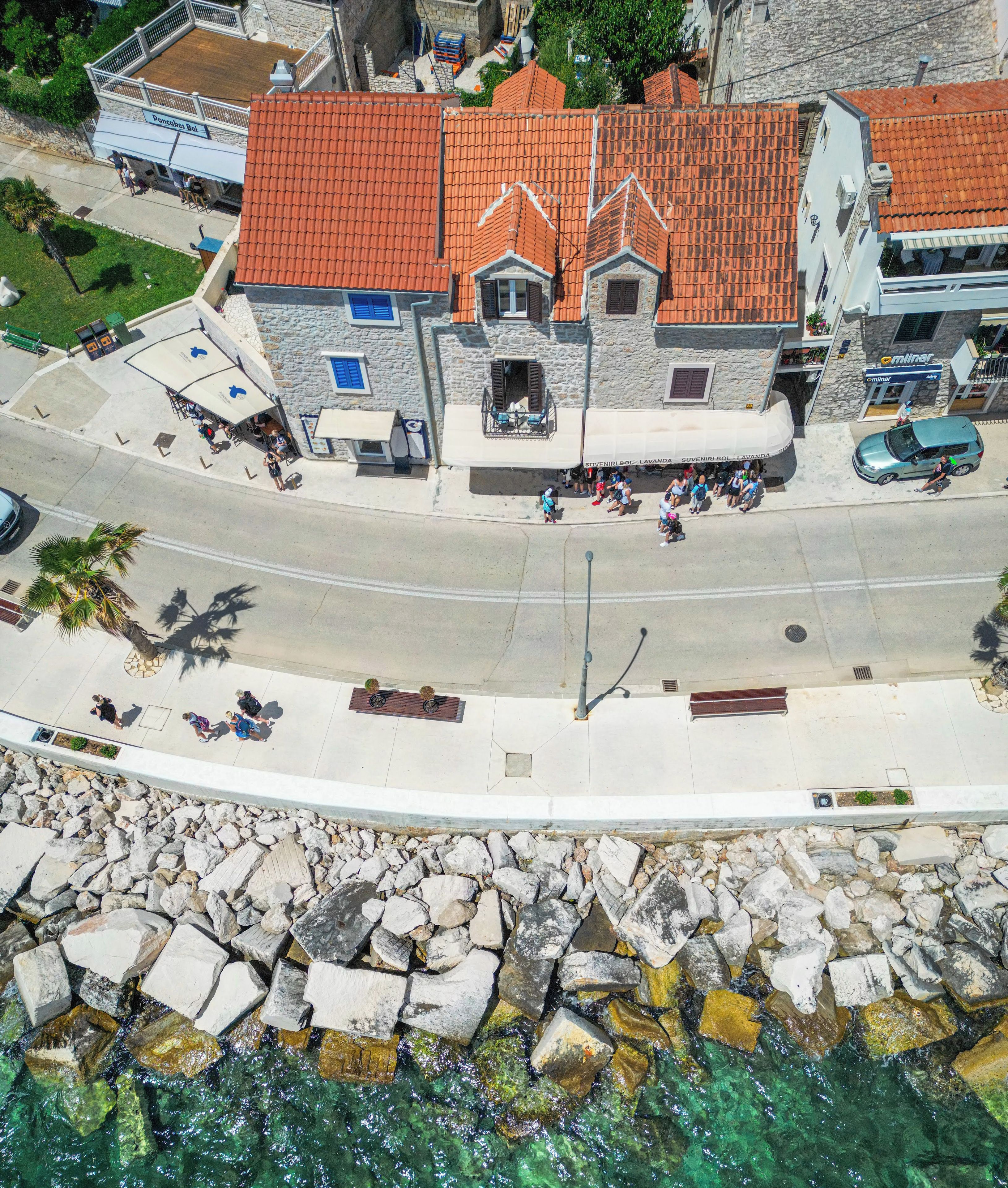 An aerial view of Riva Mare Apartments with stone buildings, a paved street, and a rocky shoreline.