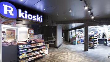 Interior de botiga amb prestatges plens d'articles diversos, un mostrador de caixa visible i un cartell que diu "R Kioski".