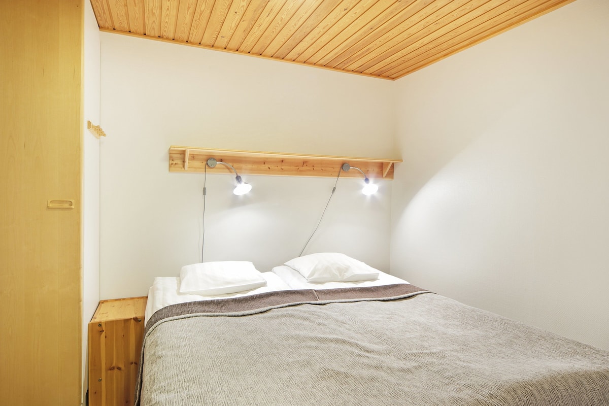 A bedroom with a wooden ceiling, a bed with white pillows and a gray blanket, and a wooden shelf with two lights.