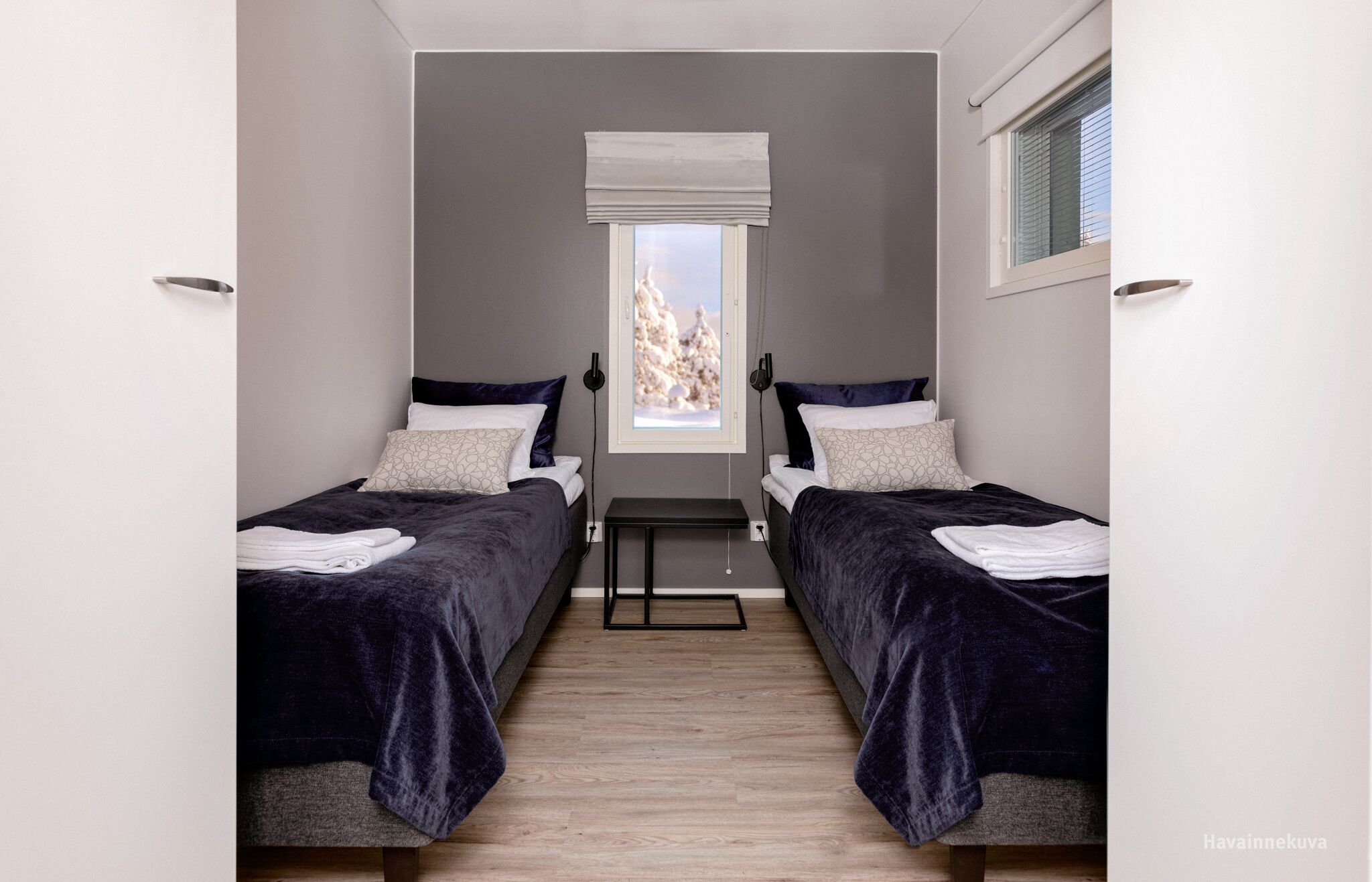 Un dormitorio moderno con dos camas, una mesita y una ventana con vistas a  árboles cubiertos de nieve.