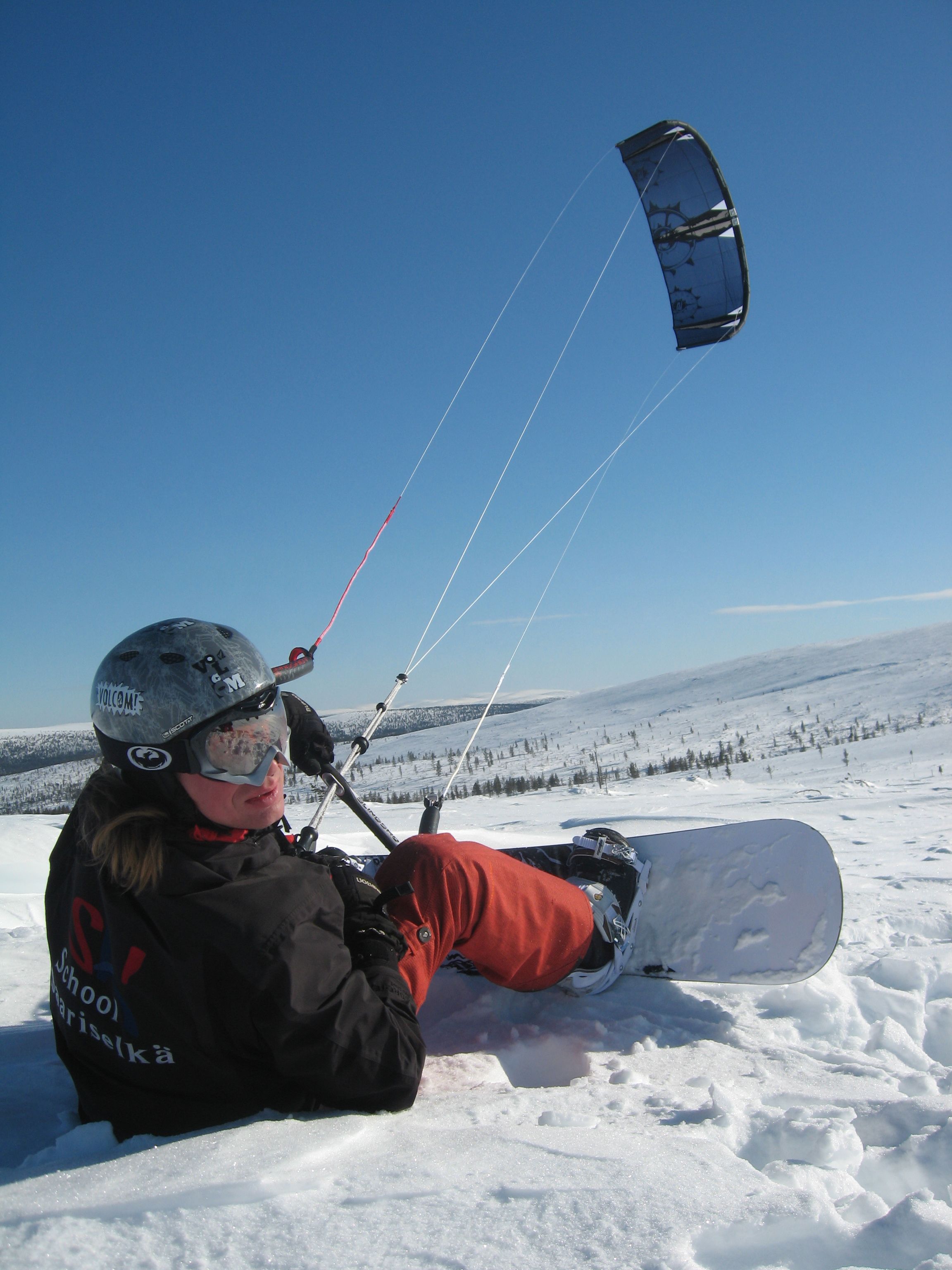 A person sitting on a snow-covered slope, wearing a helmet and holding a kite, with a clear sky and snow-covered landscape in the background.