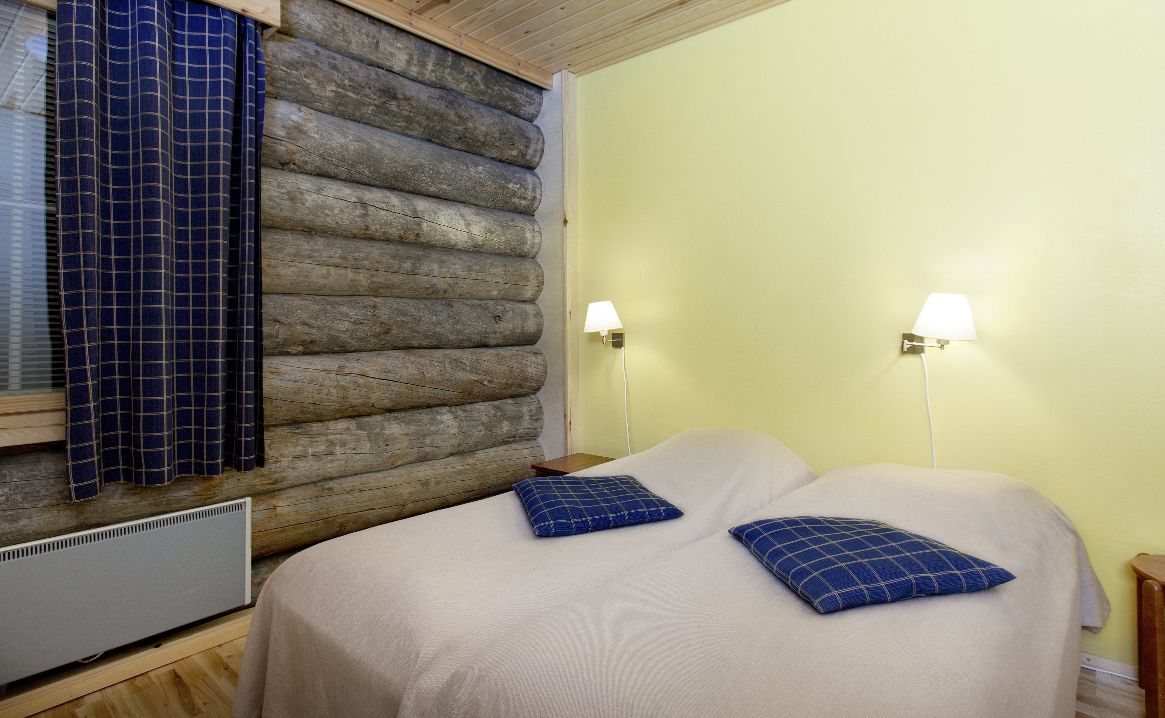 A room with a log wall, two bedside tables with lamps, a bed with two pillows, and a window with curtains.