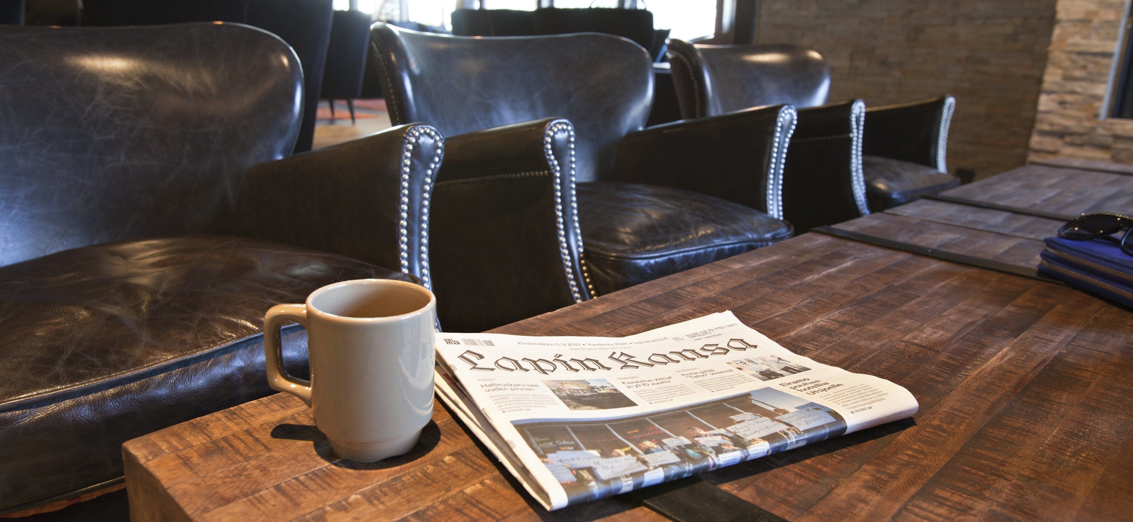 Ein Holztisch mit einer Zeitung, einer weißen Tasse und schwarzen Ledersesseln.