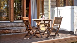 Um deck de madeira com mesa e cadeiras, janelas amplas com cortinas e fachada de cabana de tronco.