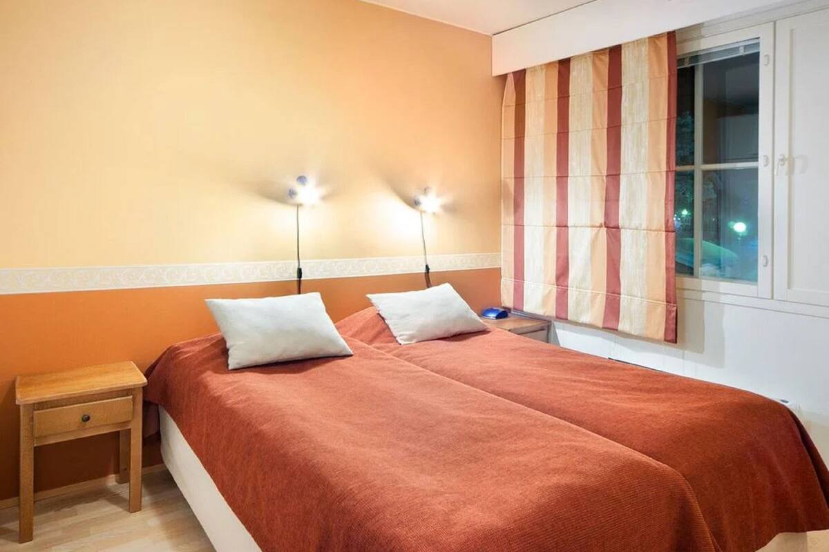 A double bed with two pillows, a wooden nightstand with a drawer, and a window with striped curtains.