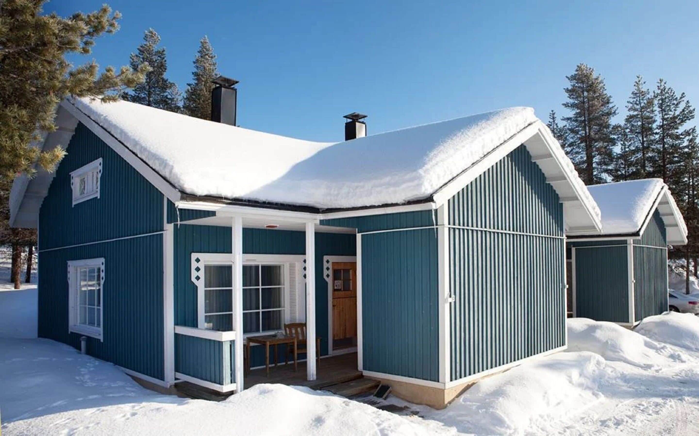 A blue cabin with a snow-covered roof, a porch, and a chimney.